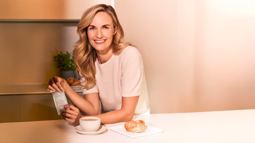 A smiling woman with long blonde hair sits at a table, holding a packet labeled "Kollagen Pulver," with a coffee cup and pastry nearby.