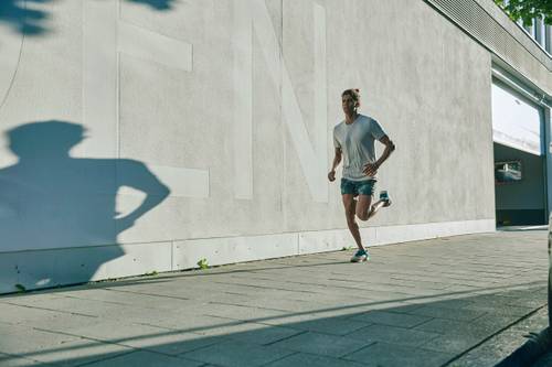A man jogs on a sunny pavement next to a large white wall, casting a shadow.