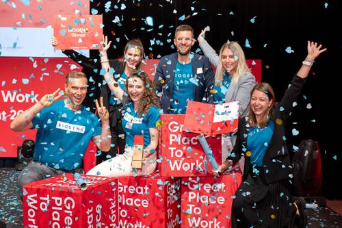 Eine Gruppe von Menschen feiert mit Konfetti und hält ein Schild mit der Aufschrift „Great Place to Work“ hoch, umgeben von roten Würfeln.