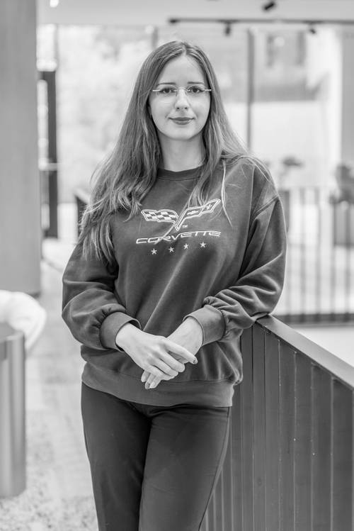 Eine Frau mit langen Haaren und Brille, die ein Corvette-Sweatshirt trägt, steht mit verschränkten Händen in einem Raum und schaut in die Kamera. Schwarz-Weiß-Foto.