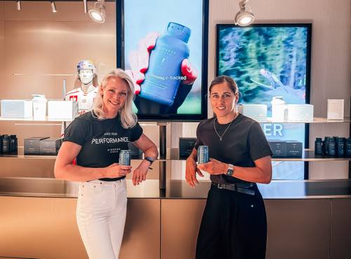 Zwei lächelnde Frauen, die Dosen in der Hand halten, stehen in einem Laden, im Hintergrund sind Produkte und digitale Bildschirme zu sehen.