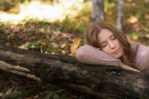Woman with red hair in a pink jumper, resting her head peacefully on a moss-covered tree trunk in a sun-drenched forest landscape.