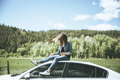 Frau sitzt auf dem Dach eines weißen Autos, umgeben von Grün und Bäumen, mit einem klaren blauen Himmel im Hintergrund.