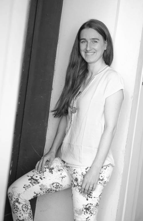 Frau lächelt, steht in einer Türöffnung, trägt eine helle Bluse und eine geblümte Hose. Schwarz-Weiß-Foto.