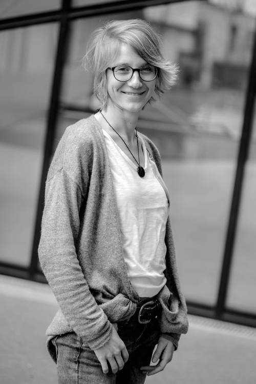 Frau mit kurzen Haaren und Brille, lächelnd, trägt eine Strickjacke und Jeans. Sie steht vor einem modernen Glasgebäude. Schwarz-Weiß-Foto.
