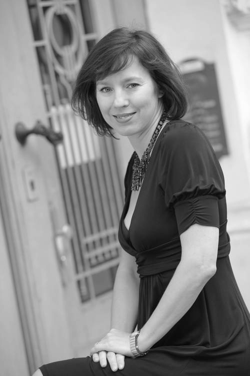 Frau mit kurzen dunklen Haaren in einem schwarzen Kleid, die auf einer Treppe sitzt, in die Kamera lächelt und sich in der Nähe einer verzierten Tür befindet. Das Foto ist in Schwarz-Weiß.