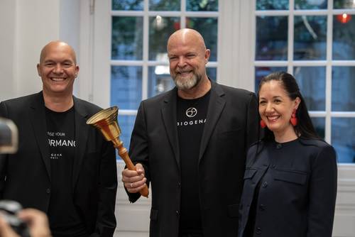 Drei Personen stehen lächelnd bei einer Veranstaltung zusammen; der Mann in der Mitte hält eine goldene Glocke in der Hand.