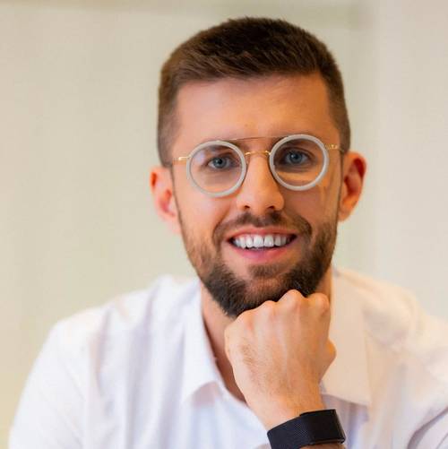 Doktor Patrick Bantsich, lächelnd mit der Hand am Kinn, mit kurzem Haar und Bart, trägt runde Brille und ein weißes Hemd.
