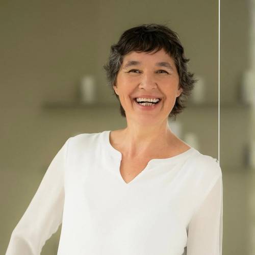 BIOGENA Wissenschafts- und Gesundheitsexpertin, Lisa Ressi, lächelnd mit kurzen dunklen Haaren, trägt eine weiße Bluse, steht in einem weich beleuchteten Raum mit verschwommenem Hintergrund.