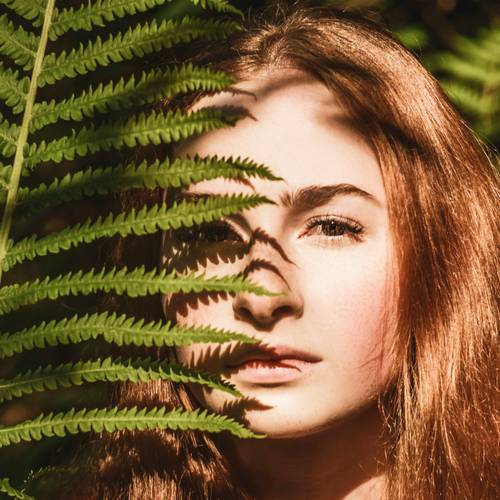 Eine Frau mit schöner Haut und langen, glänzenden roten Haaren, teilweise verdeckt von Farnblättern, Sonnenlicht wirft Schatten auf ihr Gesicht und schafft eine natürliche und ruhige Atmosphäre.