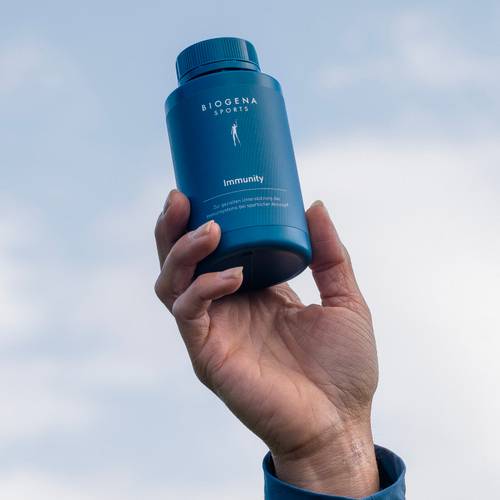 Hand, die eine blaue Flasche mit der Aufschrift „Biogena Sports Immunity” vor einem wolkigen Himmel hält.