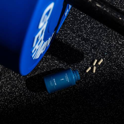 Eine blaue Flasche mit der Aufschrift „Modern Fertility“ und daneben auf einer gesprenkelten schwarzen Oberfläche verstreute Kapseln, neben einer blauen Hantelscheibe.