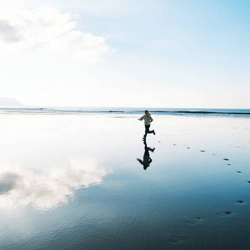 Person, die an einem reflektierenden Strand mit klarem Himmel und fernen Bergen läuft und Fußabdrücke im nassen Sand hinterlässt.