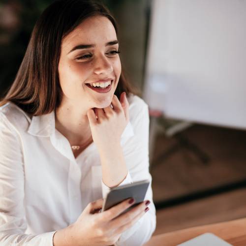 Lachende Frau in einem weißen Hemd, die die Biogena Coach-App auf ihrem Smartphone nutzt, sitzt an einem Tisch vor einem verschwommenen Hintergrund.