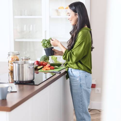 Eine vegane Frau mit langen dunklen Haaren und einem grünen Hemd kocht in einer Küche, hält einen Topf mit frischem Basilikum in der Nähe von Gemüse und einen Topf auf dem Herd. Sie lächelt, weil sie weiß, dass BIOGENA Essentials Vitamin B12-Tropfen ihre Ernährung unterstützt.
