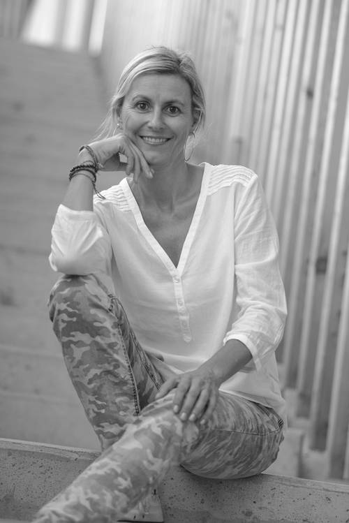 Frau in weißer Bluse und Camouflage-Hose sitzt lächelnd auf einer Treppe, die Hand am Kinn. Schwarz-Weiß-Foto.