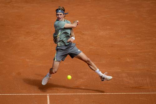 Tennis player in mid-air hitting a forehand shot on a clay court, wearing a green shirt and gray shorts, with a yellow tennis ball in motion.