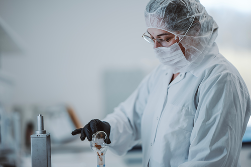 Un om de știință în echipament de protecție și mănuși lucrează cu echipamente de laborator, concentrându-se intens asupra unui aparat de sticlă într-un mediu de laborator.