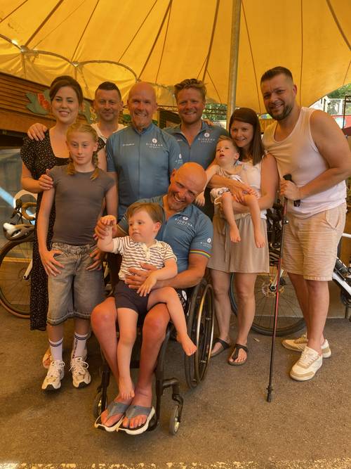 A group of people, including a man in a wheelchair and children, posing under a yellow canopy with bicycles in the background.