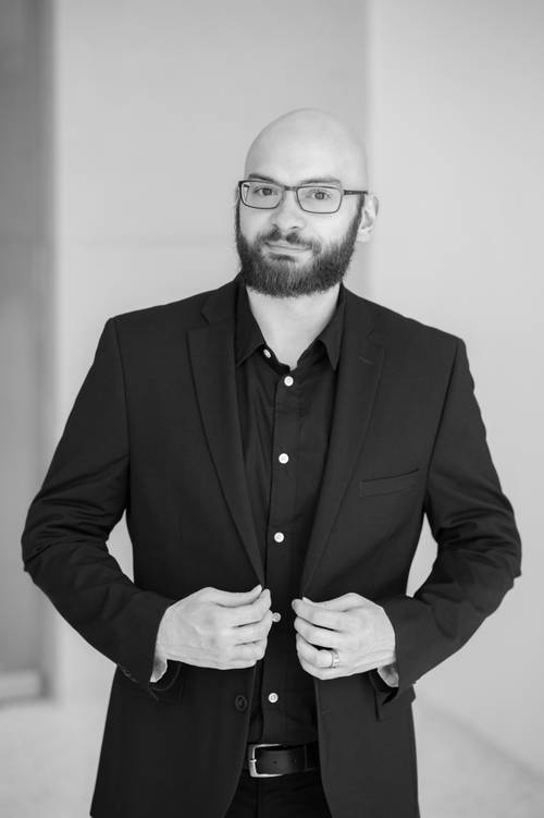 Bărbat chel, cu barbă și ochelari, îmbrăcat într-un costum negru, cu haina încheiată la gât, stând în fața unui fundal monocrom.