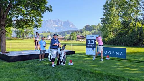 Golfer in a pink shirt tees off on a lush course with mountain views, while others watch nearby near a sign marked "13" and "BIOGENA."