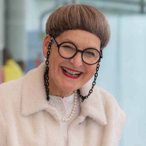Susanne "Nana" Bernegger-Flintsch lächelnd, mit kurzen Haaren, runden Brillen und weißen Mantel, trägt eine Perlenkette und eine schwarze Brillenkette.