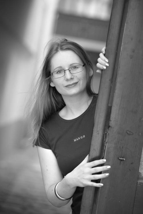 Eine Person mit Brille und schwarzem Hemd späht hinter einer Holztür hervor auf einem Schwarz-Weiß-Foto.