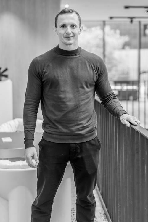 Eine Person in Pullover und Hose steht drinnen an einem Geländer, im Hintergrund sind eine moderne Inneneinrichtung und der Blick aus dem Fenster zu sehen. Schwarz-Weiß-Bild.