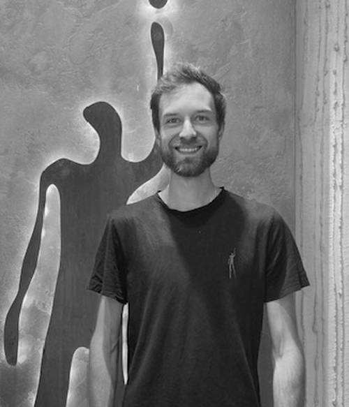 Mann lächelt vor einer Wand mit künstlerischem Silhouetten-Design und trägt ein schwarzes T-Shirt. Schwarz-Weiß-Foto.