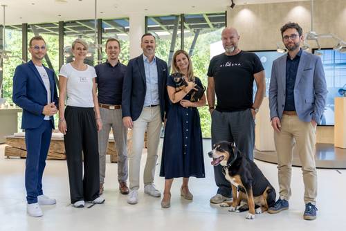 Gruppenfoto von sieben Personen und zwei Hunden in einem modernen Innenraum; eine Frau hält einen kleinen Hund, ein größerer Hund sitzt im Vordergrund.