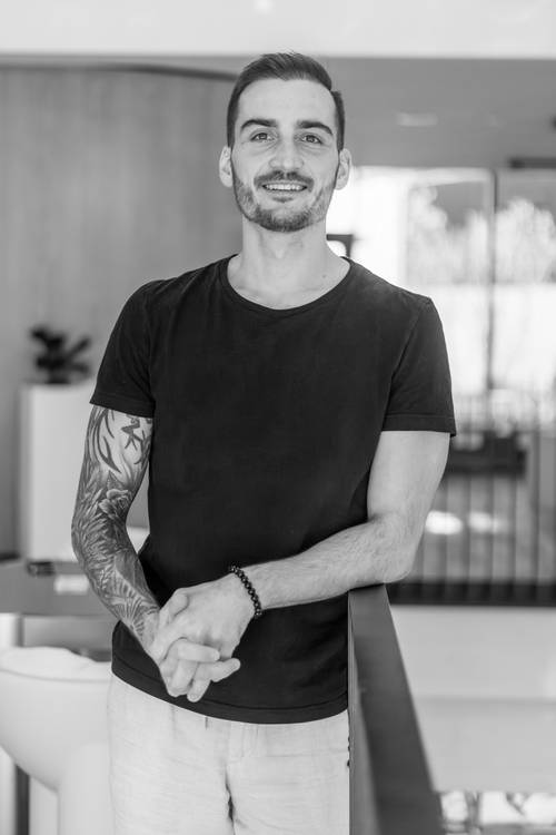Ein lächelnder Mann mit tätowiertem Arm und schwarzem T-Shirt steht in einem modernen Raum an einem Geländer. Schwarz-Weiß-Fotografie.