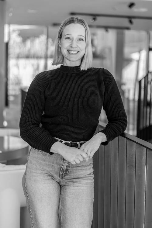 Schwarz-Weiß-Foto einer lächelnden Frau mit schulterlangem Haar in einem Pullover, die sich in einem Innenraum an ein Geländer lehnt.