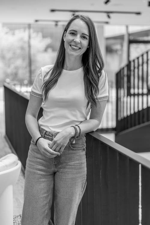 Lächelnde Frau in einem weißen Oberteil und Jeans, die sich in einer modernen, hellen Innenraumumgebung an ein Geländer lehnt.
