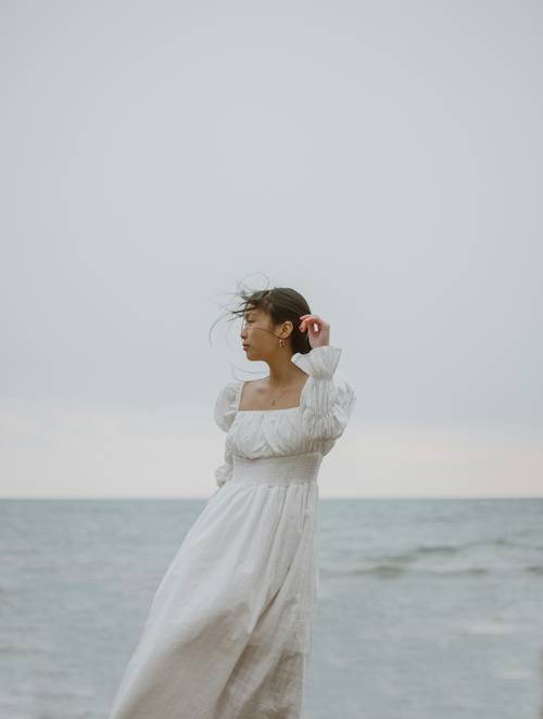 Eine Frau in einem wallenden weißen Kleid steht am Meer, während der Wind ihr Haar vor dem grauen Himmel zerzaust.