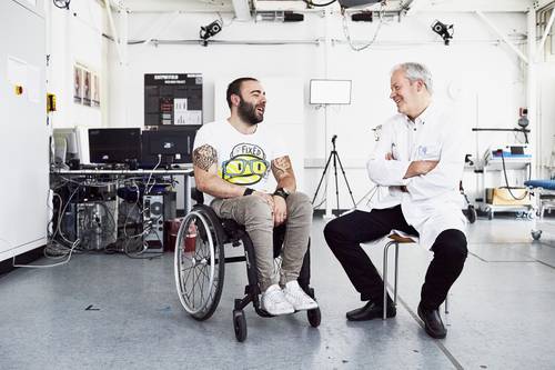 Zwei Männer in einem hellen Labor: Der eine sitzt in einem Rollstuhl und trägt Freizeitkleidung, der andere trägt einen weißen Kittel und lächelt; beide unterhalten sich.