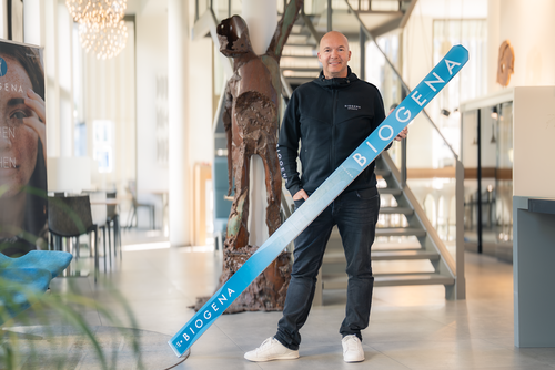 Eine Person in einer schwarzen Jacke hält einen großen blauen BIOGENA-Ski in einem modernen Büro; im Hintergrund sind eine Treppe und eine Skulptur zu sehen.