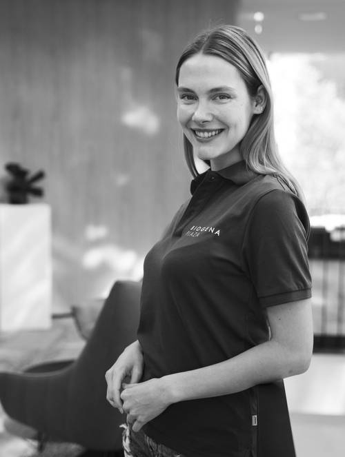 Lächelnde Frau in einem Polohemd steht in einem modernen Interieur mit sanfter Beleuchtung und unscharfem Hintergrund.
