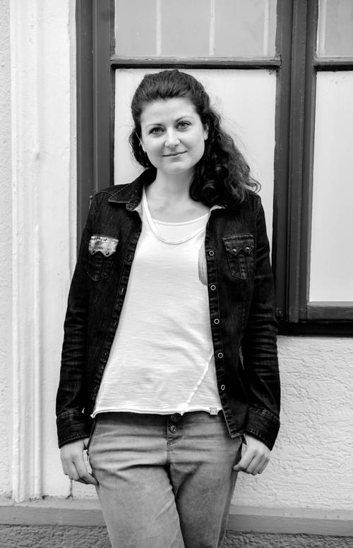 Frau in weißem Hemd und Jeansjacke steht mit neutralem Gesichtsausdruck vor einem Fenster; Schwarz-Weiß-Foto.
