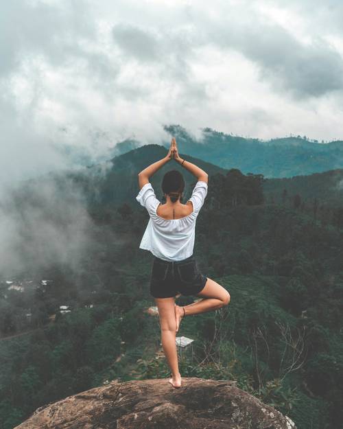 Person, die Yoga in der Baumhaltung auf einer Klippe praktiziert, umgeben von nebligen Bergen und üppigem Grün.