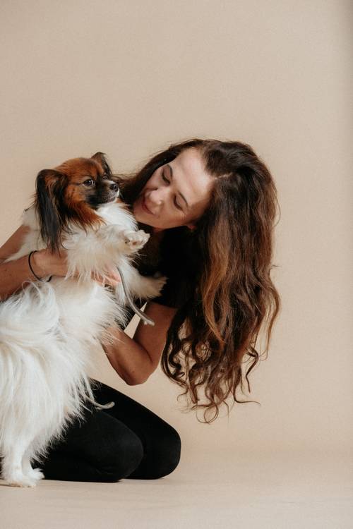 Frau mit langen Haaren kniet und hält einen flauschigen Hund im Arm. Beide schauen sich liebevoll an, vor einem beigen Hintergrund.