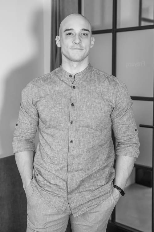 Eine Person in einem Hemd mit Knöpfen steht selbstbewusst mit den Händen in den Taschen und einem leichten Lächeln in einem modernen Innenraum. Schwarz-Weiß-Foto.