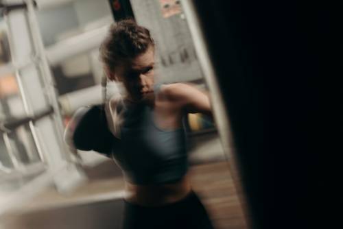 Frau mit geflochtenen Haaren in einem Sport-Top schlägt mit einer Faust auf einen Boxsack in einer Bewegungsunschärfe-Aufnahme in einem Fitnessstudio.