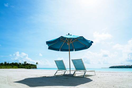 Two deck chairs under a blue sun umbrella on a sandy beach, with a clear sky and palm trees in the distant background.