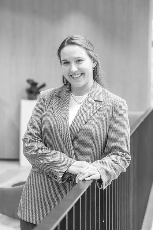 Lächelnde Frau in einem karierten Blazer lehnt sich an ein Geländer in einem Innenraum, mit unscharfem Hintergrund und einer Topfpflanze. Schwarz-Weiß-Foto.
