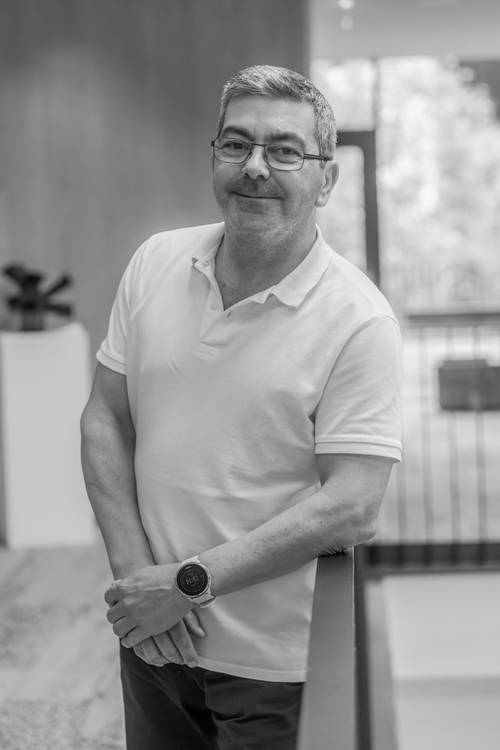 Ein Mann in einem weißen Polohemd und mit Brille lehnt lächelnd an einem Geländer in einem Innenraum. Im Hintergrund sind große Fenster und verschwommene Einrichtungsgegenstände zu sehen. Schwarz-Weiß-Foto.