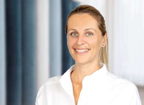 Smiling woman with blonde hair in a white shirt stands in a bright room with a blurred background of windows and blue panels.