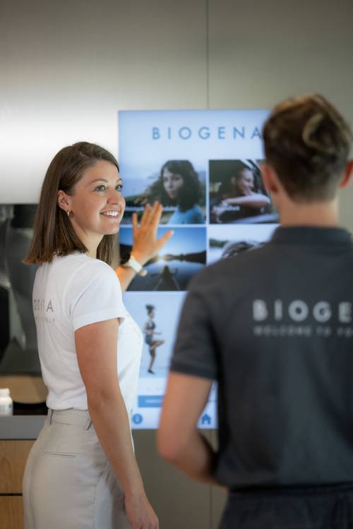 Zwei Personen in gebrandeten Shirts stehen neben einem BIOGENA-Display. Eine davon zeigt auf die Bilder auf dem Bildschirm, lächelt und führt ein Gespräch.