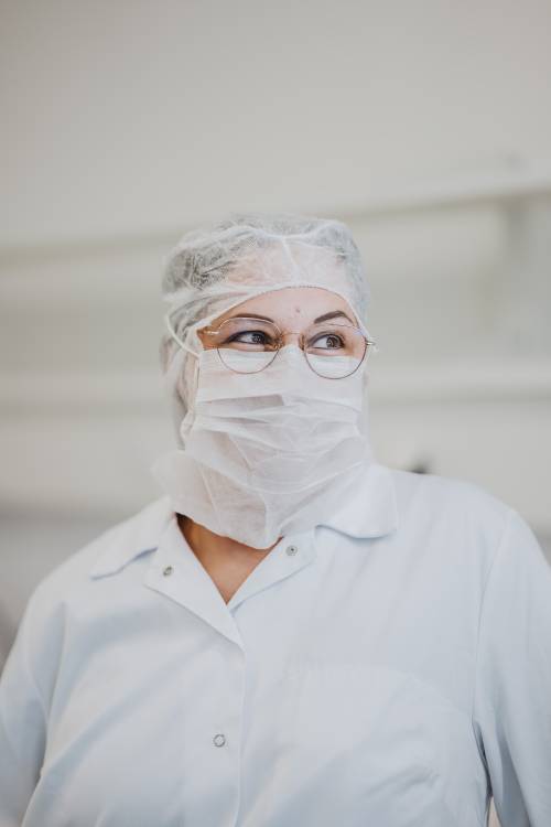 Persoană în halat de laborator, plasă de păr și mască, purtând ochelari, privind într-o parte cu o expresie concentrată într-un mediu luminos de laborator.