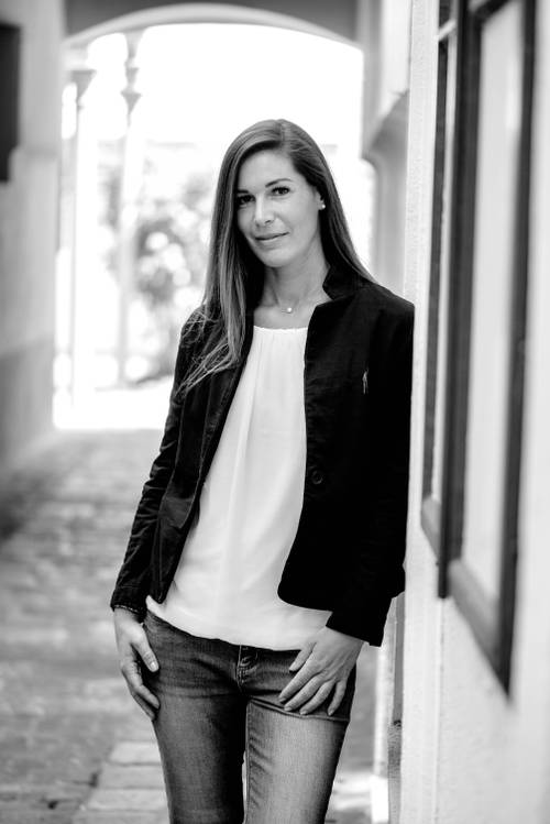Schwarz-Weiß-Foto einer Frau mit langen Haaren, die eine Jacke und Jeans trägt und an einer Wand in einer engen Gasse lehnt.