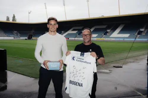 Zwei Leute stehen in einem Stadion, einer hält ein signiertes Fußballtrikot in der Hand, der andere eine Plakette, und beide lächeln in die Kamera.